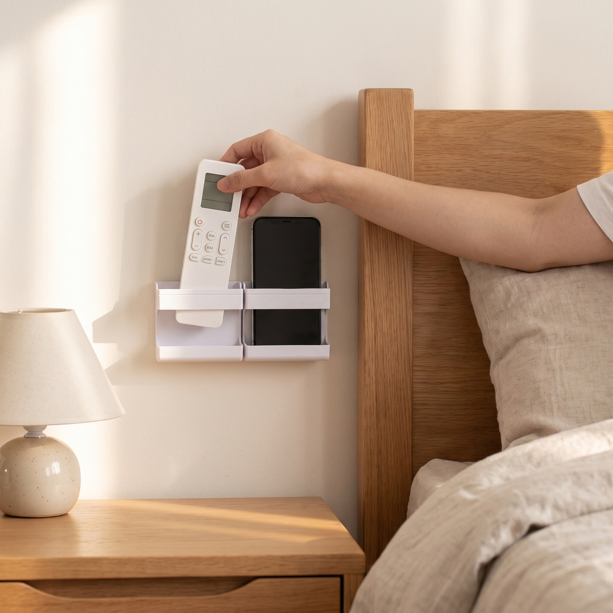 Adhesive wall caddy beside a bed holding a remote and a phone, hand lifting the remote out