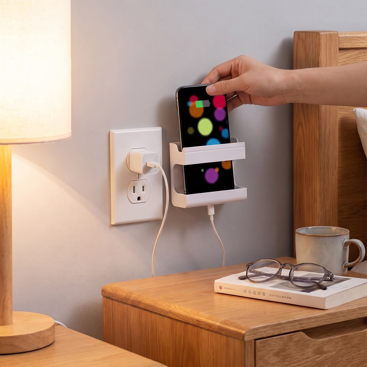Wall-mount holder beside a bed holding a charging phone above a wall outlet